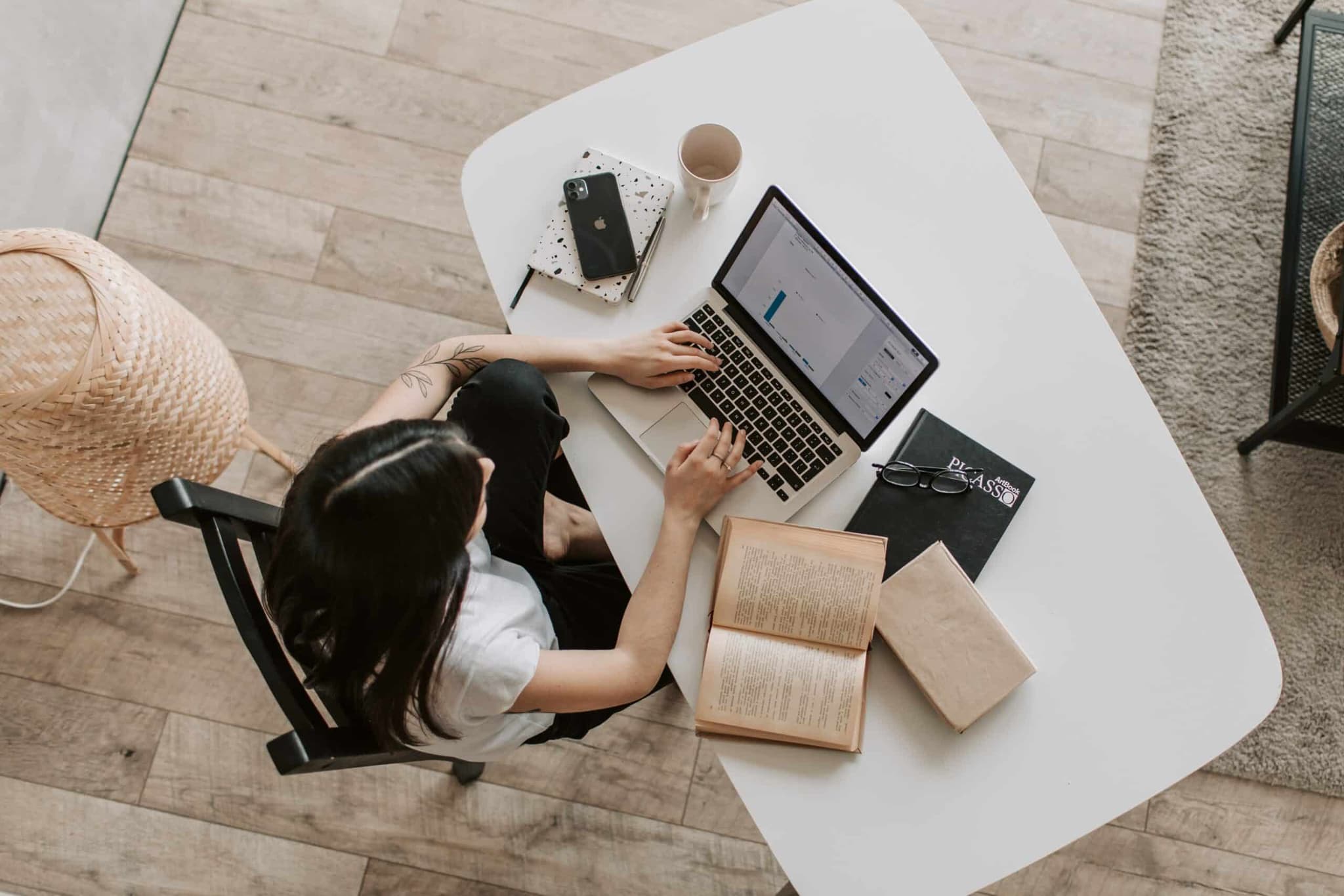 From a top-down view, a young woman sitting at a desk with her laptop open in front of her. She appears to be browsing the internet and is surrounded by books, papers, and other items.