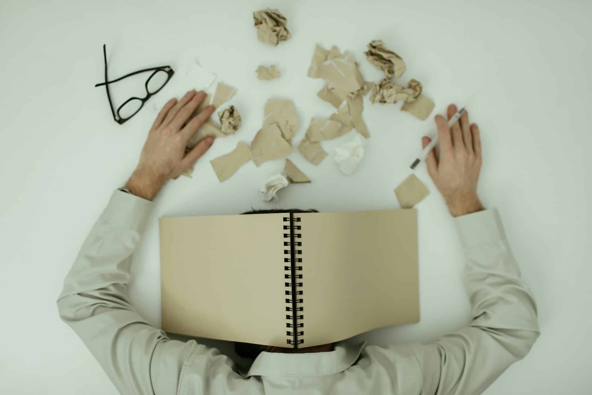 A person resting his head on a table looking distressed. His head covered with a notebook and scattered papers on the table.