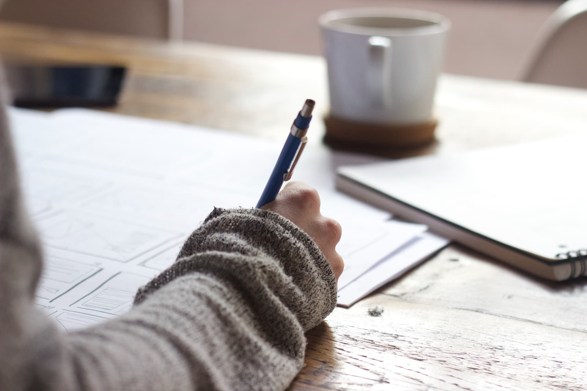 A hand writes on a sheet of paper with a cup of coffee by its side.