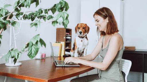 A woman looking at her laptop with her dog at the table.