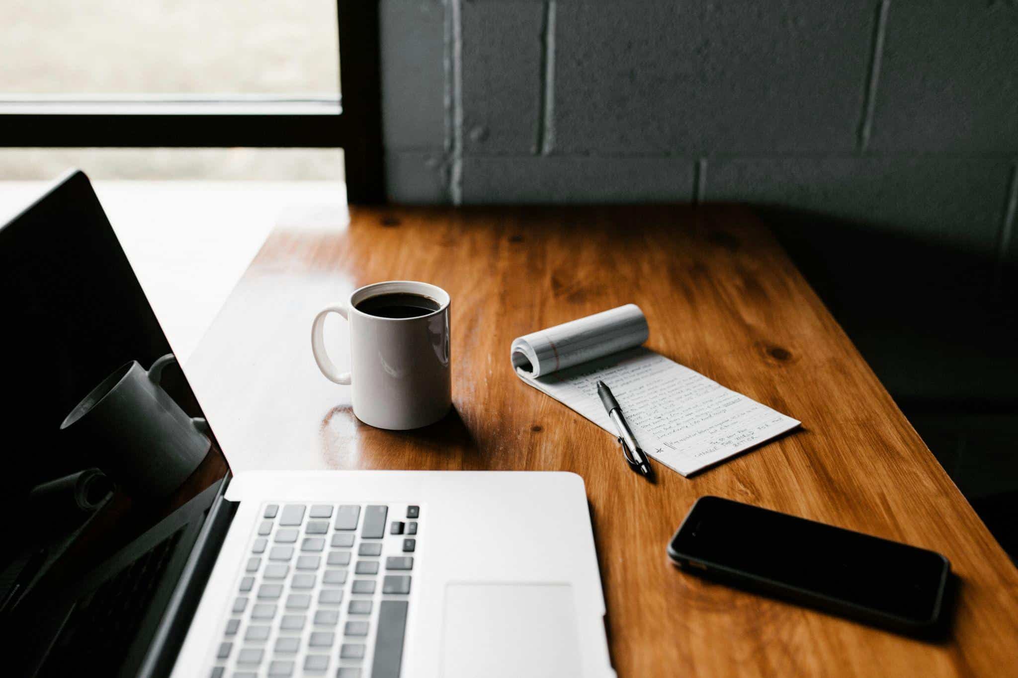 Desk by a window with a laptop, notebook, coffee cup, and smartphone